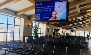 This image features a look at the airport terminal with seating and a large monitor with Kevin Costner and message that reads "Download the Autio App to start exploring Santa Barbara & get your first 5 audio stories free"