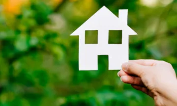 This image is a universal housing image showing a person holding a cutout of a typical house against a green grass backbround