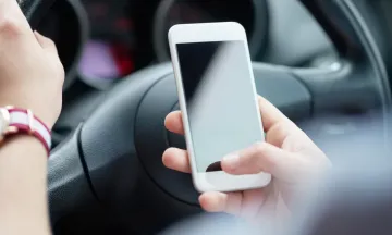 Image shows a person holding a phone in one hand and the steering wheel with the other hand