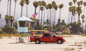 Image of East Beach, showing a row of palm trees lining the sand with a blue Lifeguard stand and red Lifeguard truck