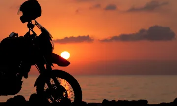 Image shows a parked motorcycle against a sunset background and the ocean. A helmet rests on the handlebars