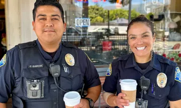 Two officers, a man and a woman, smile into the camera and hold to go cups of coffee
