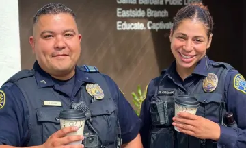 Two officers, a man and a woman, smile into the camera and hold to go cups of coffee