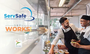 Two men in cook uniforms talk and smile in a kitchen