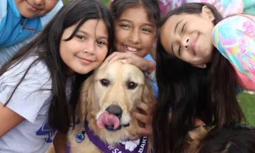 A group of children smile with a golden colored dog in a Therapy Dogs of Santa Barbara bandana 