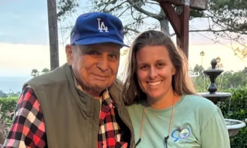 LeRoy Villa on the left in a red black and white checkered flannel, a jacket, and blue LA hat, standing next to Jaime Diamond in a green long sleeve shirt with long brown hair; both smiling.