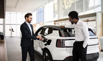 Dealer sells electric SUV to businessman in car dealership. They discuss vehicle features. Business environment and modern electric vehicle concept.