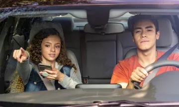 Two teens in a car, one driving with eyes on the road, the other holding the phone and navigating.