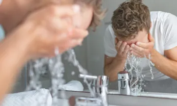 A man is washing his face in front of a sink and a mirror.