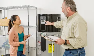 A contractor explains the settings on a heat pump water heater to a homeowner, who is holding informational paperwork. 