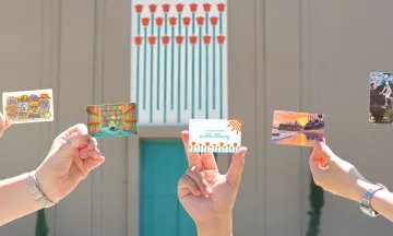 Hands holding library cards in front of the Sunburst Over California Poppies mural.