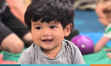 Smiling toddler in a gray shirt with another child and toys in the background.