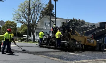 Workers wearing bright yellow shirts paving a residential street with heavy machinery.