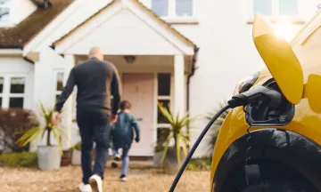 Stock image of an electric vehicle charging as family walks into home in the background