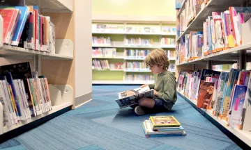 Child reading a book while sitting on a blue carpeted floor in a library aisle.