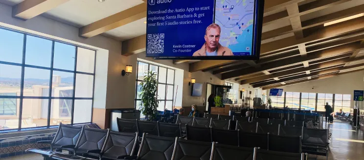 This image features a look at the airport terminal with seating and a large monitor with Kevin Costner and message that reads "Download the Autio App to start exploring Santa Barbara & get your first 5 audio stories free"