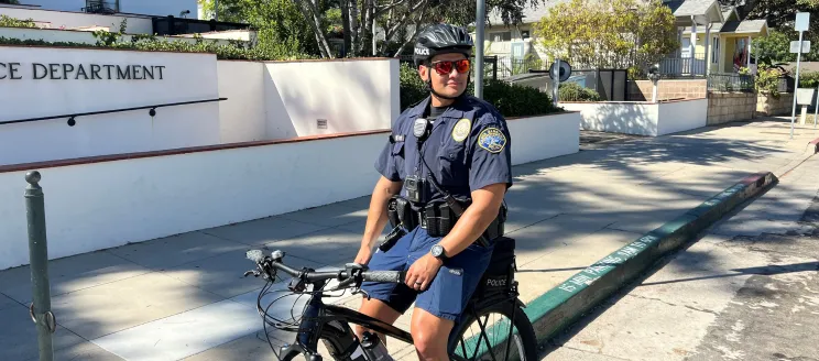 Police Department Rolls out E-Bikes to Their Fleet, image shows officer in front of Police Station on e-bike
