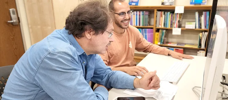 Two adults sitting at a workspace together looking at a computer 