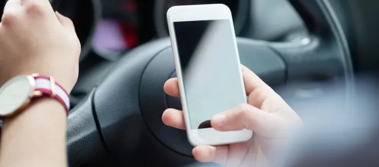 Image shows a person holding a phone in one hand and the steering wheel with the other hand