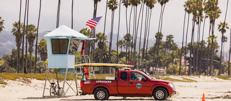 Image of East Beach, showing a row of palm trees lining the sand with a blue Lifeguard stand and red Lifeguard truck