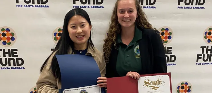 Two teen girls pose for a photo with certificates in front of a step and repeat