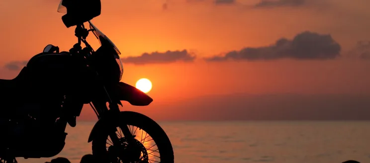 Image shows a parked motorcycle against a sunset background and the ocean. A helmet rests on the handlebars