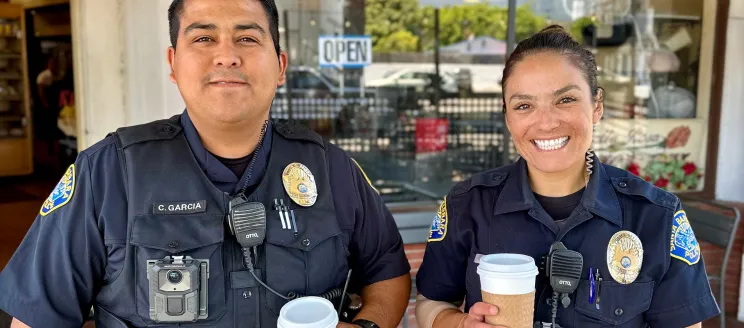 Two officers, a man and a woman, smile into the camera and hold to go cups of coffee