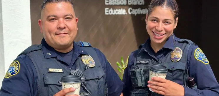 Two officers, a man and a woman, smile into the camera and hold to go cups of coffee