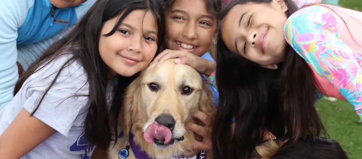 A group of children smile with a golden colored dog in a Therapy Dogs of Santa Barbara bandana 