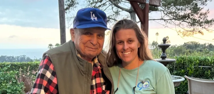 LeRoy Villa on the left in a red black and white checkered flannel, a jacket, and blue LA hat, standing next to Jaime Diamond in a green long sleeve shirt with long brown hair; both smiling.