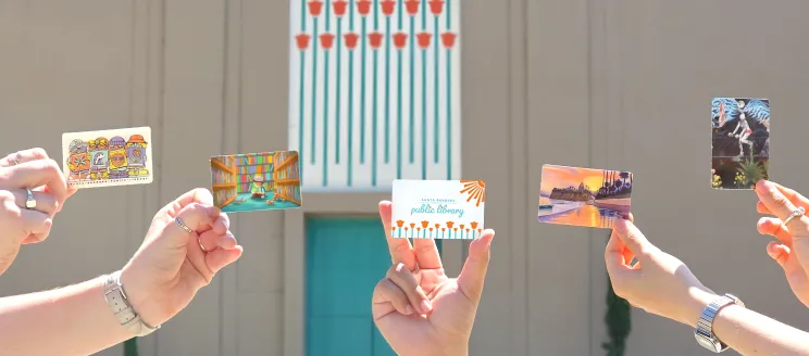 Hands holding library cards in front of the Sunburst Over California Poppies mural.