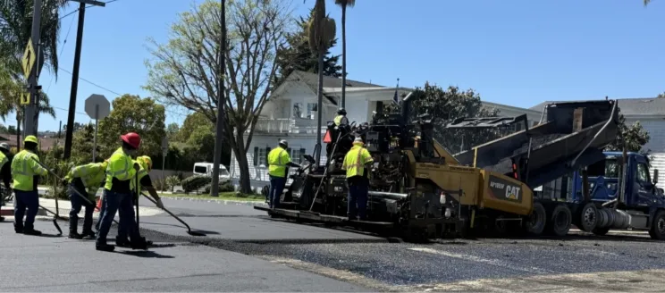 Workers wearing bright yellow shirts paving a residential street with heavy machinery.