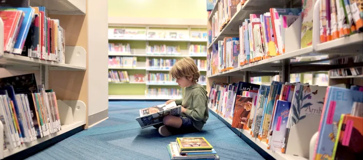 Child reading a book while sitting on a blue carpeted floor in a library aisle.