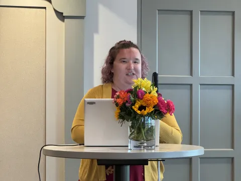 Image of Chelsea Crabill-Hauptli standing at a table with a laptop and beautiful bouquet of flowers speaking to an audience