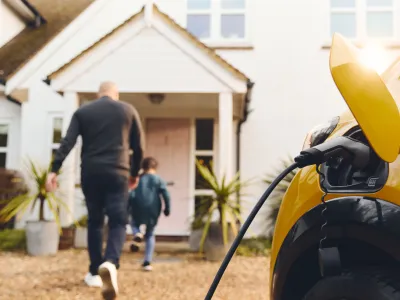 Stock image of an electric vehicle charging as family walks into home in the background